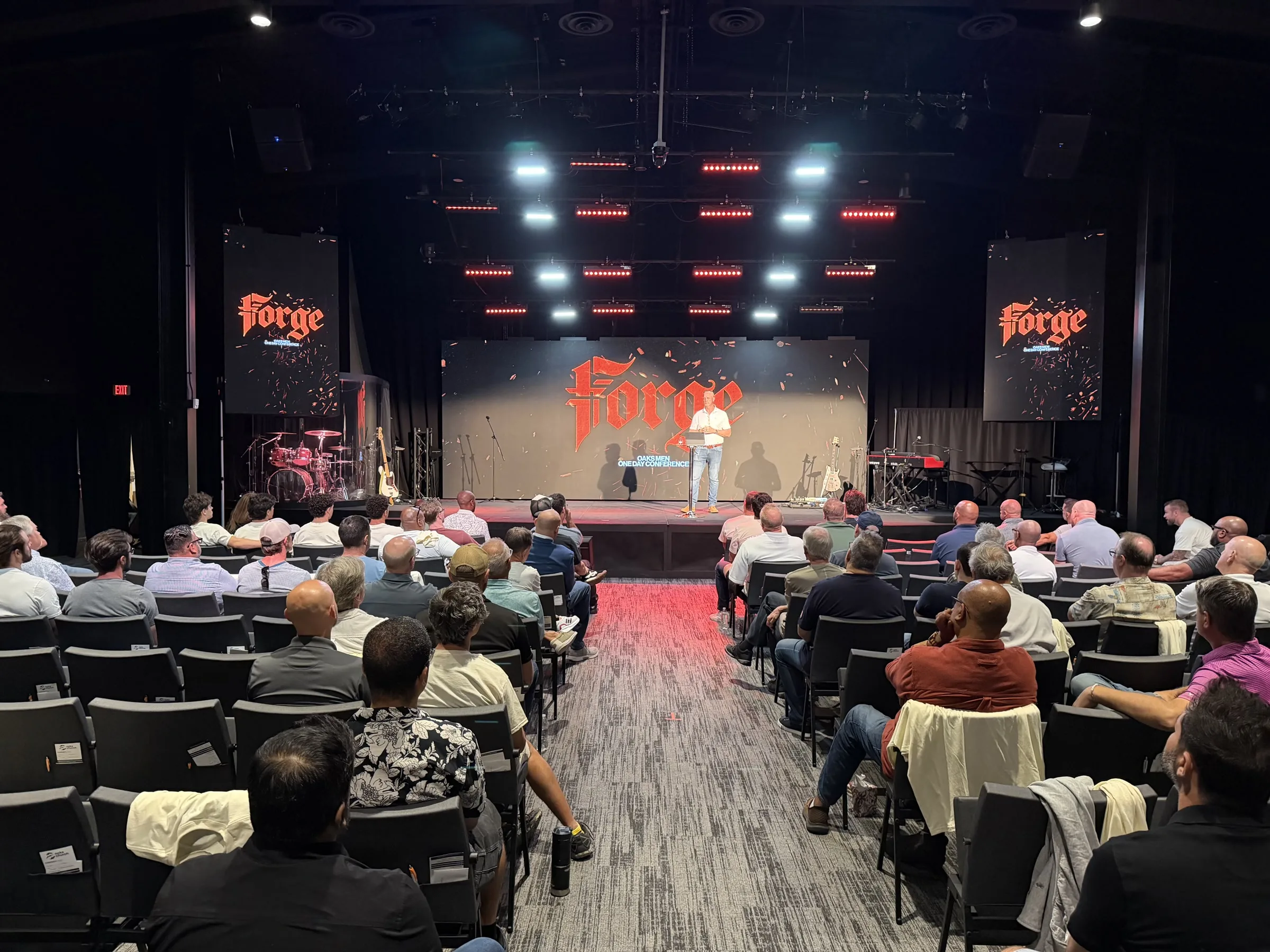 A wide view of a speaker and men gathered in the Forge One Day auditorium