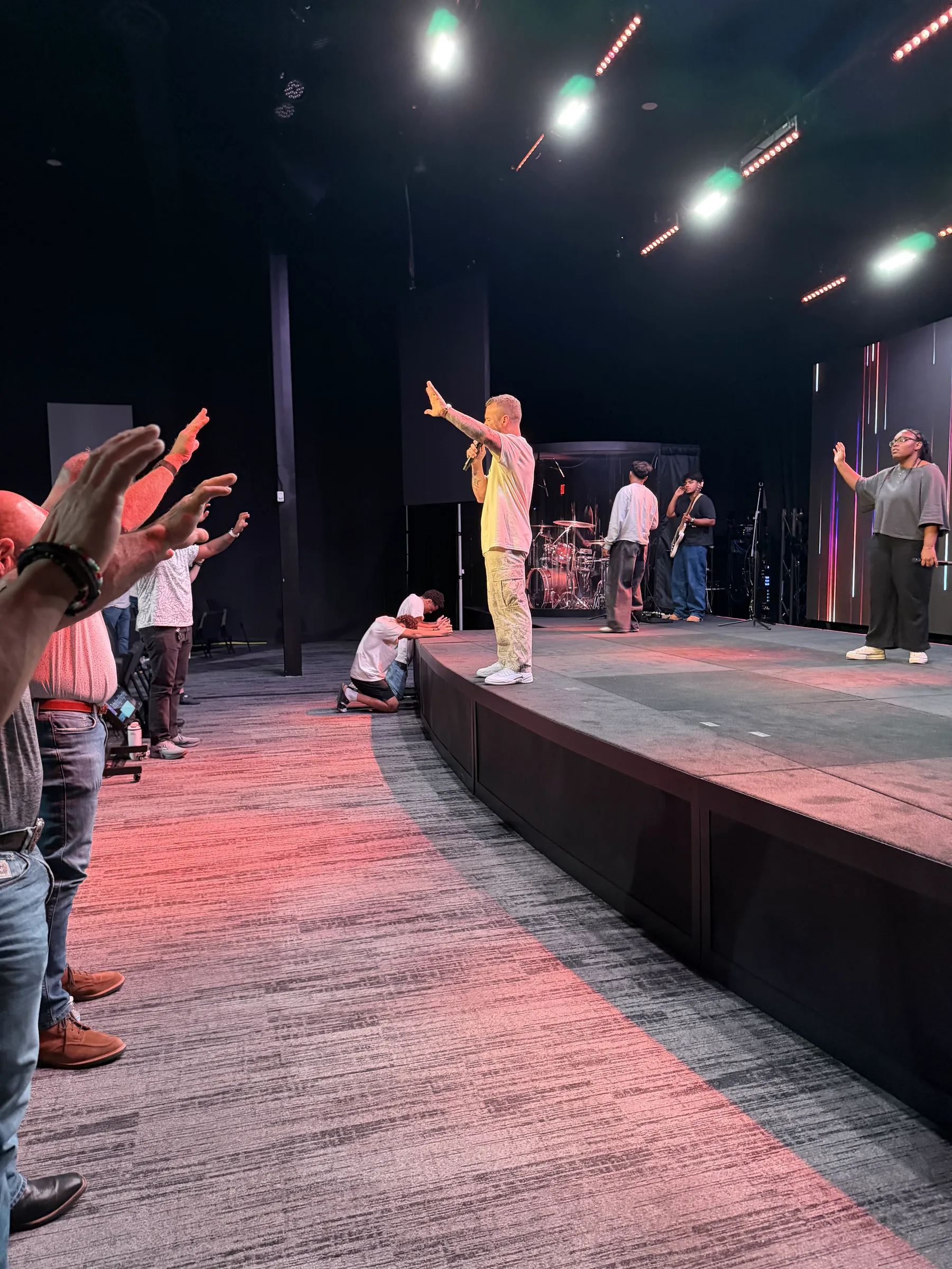 A worship leader prays from the Forge One Day stage as men reach forward and one kneels at the front